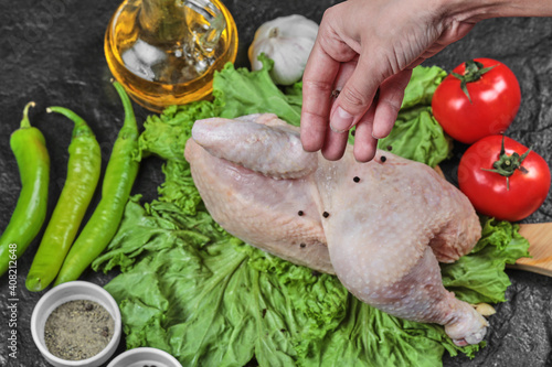 Woman adds spices to raw chicken with bunch of vegetables on dark background