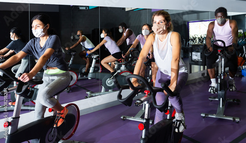 Multiethnic group of young adults in protective face masks training on cycling machines in gym during coronavirus pandemic