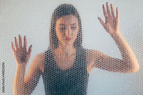 Woman protection. Defocused portrait. Self isolation. Female discrimination. Hopeless lady in black top trapped behind transparent bubble wrap texture wall on light background out of focus.