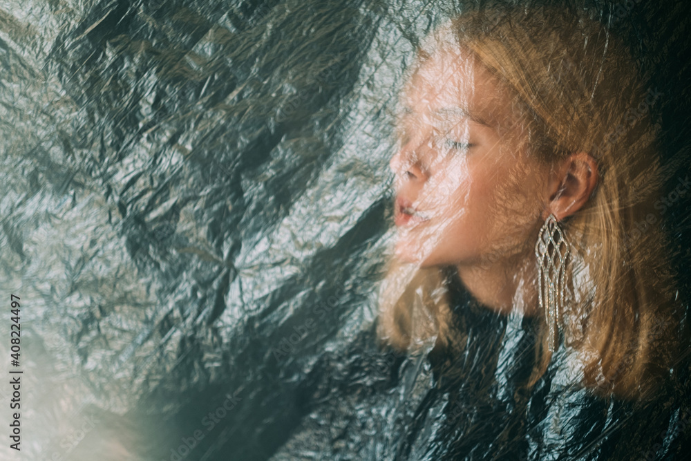 Female loneliness. Defocused art portrait. Disappointment sorrow. Self isolation. Tranquil hurt blonde woman face behind wrinkled texture transparent polyethylene film on dark background out of focus.