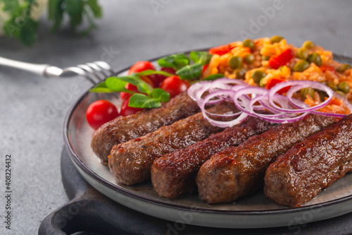 Close-up of beef kofta or cevapcici with rice and vegetables, decorated with red onion. On grey table.