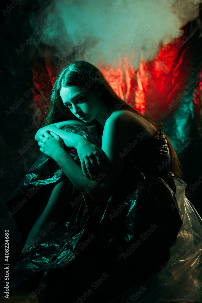 Woman abuse. Female rights. Harassment pressure. Art portrait of hurt depressed lady wearing black garbage bag dress in blue neon light on abstract dark red glowing volcano burn smoke background.