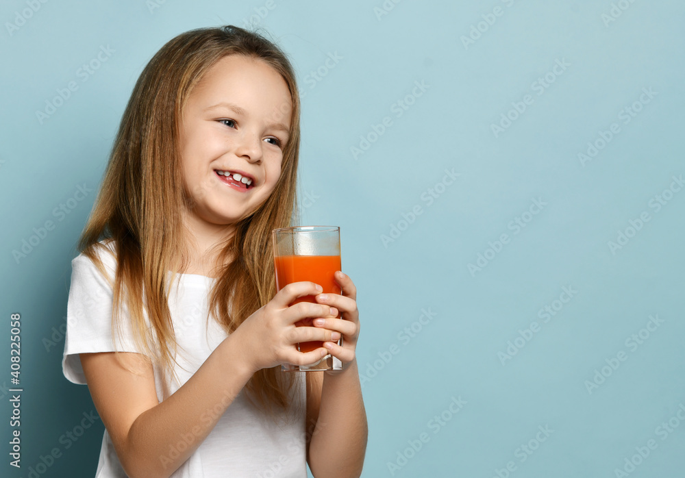 Portrait of cheerful happy smiling cute kid girl in white t-shirt standing with glass of fresh fruit juice and looking at copy space over blue background. Happy childhood, healthy lifestyle concept Portrait of cheerful happy smiling cute kid girl in white t-shirt standing with glass of fresh fruit juice and looking at copy space over blue background. Happy childhood, healthy lifestyle concept