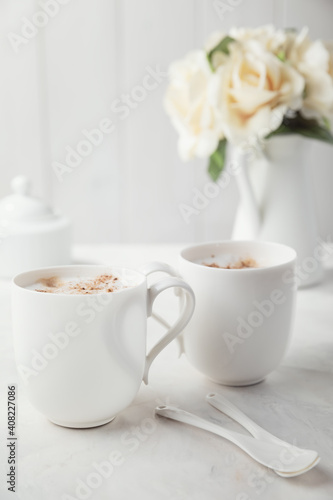 Two white porcelain cups of coffee. Bouquet of delicate cream flowers in the background. Romantic morning for two.
