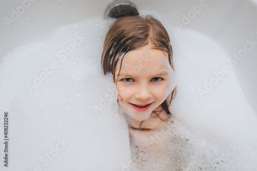 A small, smiling, cute red-haired girl with long hair, a child bathes, lying in a white bath with foam from soap and shampoo. Model photography.