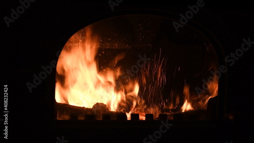 Blurry background of retro fireplace with burning firewoods and glowing flame inside with bright spark traces. Cozy warm light in night darkness. Romantic backdrop for winter season