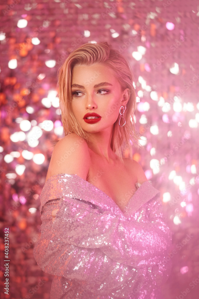 Fashionable partly defocused blurred studio portrait of young sexy blonde woman with makeup and middle length straight hair in shining silver dress against glamor sequins background. Pink light