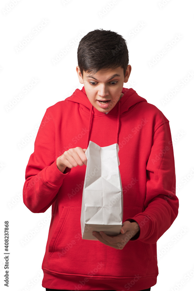 Surprised overjoyed teenage boy opening white paper bag looking inside with facial expression. Attractive caucasian teenager dressed in red hoodie holding package isolated on white. Surprised overjoyed teenage boy opening white paper bag looking inside with facial expression. Attractive caucasian teenager dressed in red hoodie holding package isolated on white.