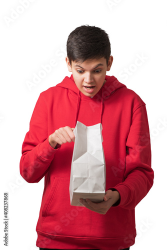 Obraz na płótnie Surprised overjoyed teenage boy opening white paper bag looking inside with facial expression. Attractive caucasian teenager dressed in red hoodie holding package isolated on white. w salonie