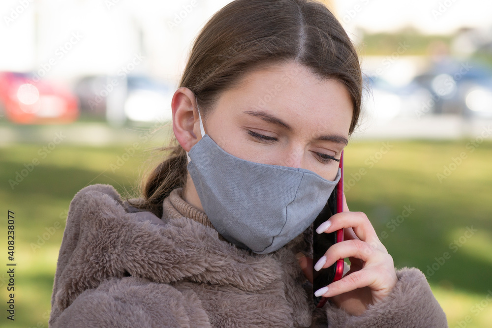 Close up of young pretty Caucasian woman standing in city in face mask and talking on smartphone. Portrait of beautiful female calling and chatting on cellphone outdoors in quarantine. Leisure concept Close up of young pretty Caucasian woman standing in city in face mask and talking on smartphone. Portrait of beautiful female calling and chatting on cellphone outdoors in quarantine. Leisure concept