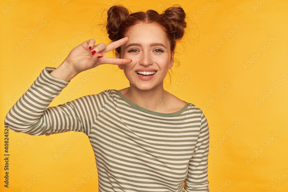 Young lady, pretty, cheerful ginger woman with two buns. Wearing striped blouse and showing peace sign over her eye, smiling. Watching at the camera isolated over yellow background Young lady, pretty, cheerful ginger woman with two buns. Wearing striped blouse and showing peace sign over her eye, smiling. Watching at the camera isolated over yellow background