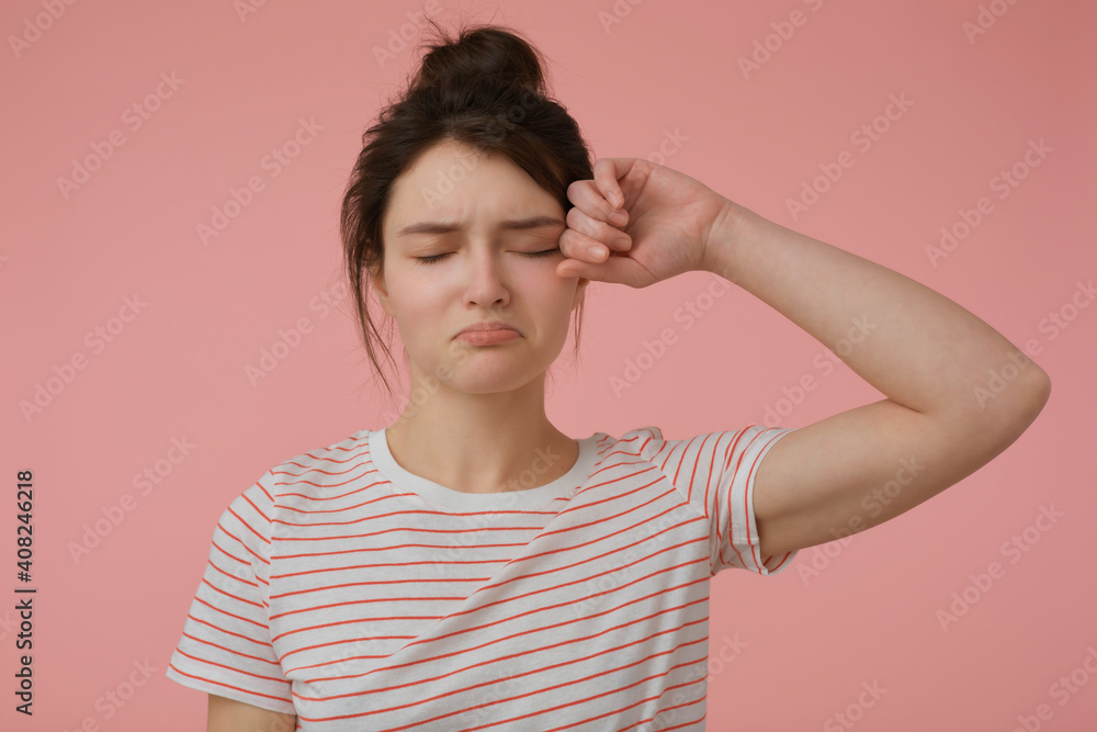 Portrait of attractive, upset girl with brunette hair and bun. Wearing t-shirt with red strips and rub an eye. Emotional concept. Stand isolated over pastel pink background Portrait of attractive, upset girl with brunette hair and bun. Wearing t-shirt with red strips and rub an eye. Emotional concept. Stand isolated over pastel pink background