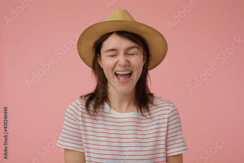 Teenage girl, happy looking woman with long brunette hair. Wearing t-shirt with red strips and hat. Emotional concept. Screams with closed eyes. Stand isolated over pastel pink background