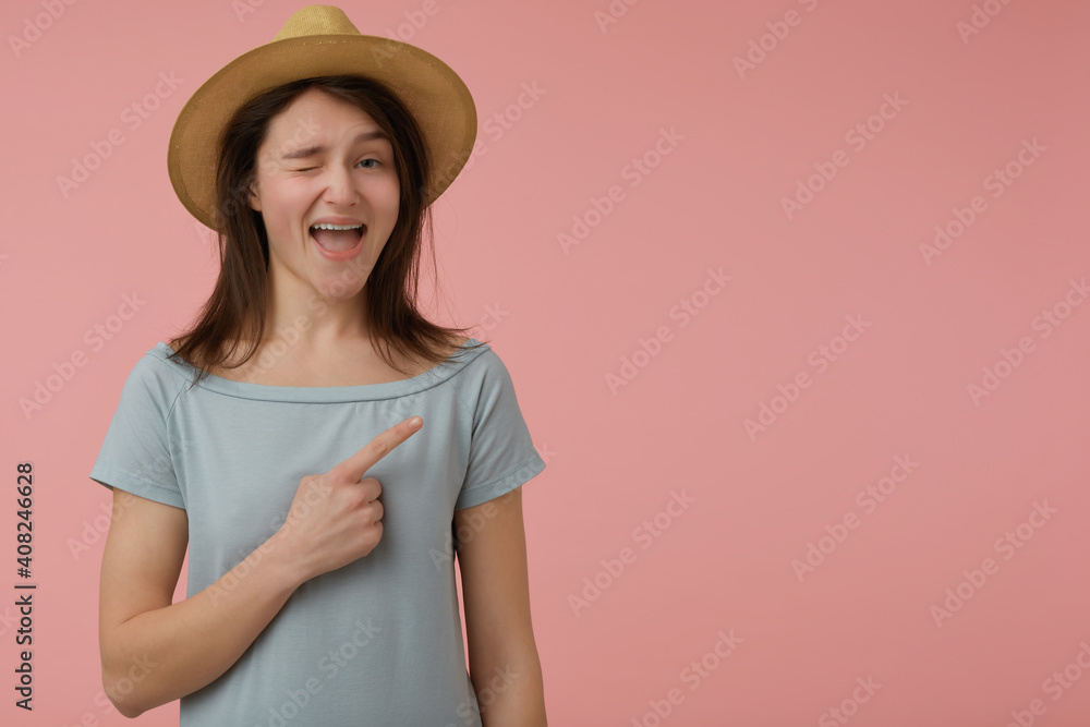 Portrait of attractive, adult girl with long brunette hair. Wearing blueish t-shirt and hat. Wink, smiling at the camera and pointing to the right at copy space over pastel pink background Portrait of attractive, adult girl with long brunette hair. Wearing blueish t-shirt and hat. Wink, smiling at the camera and pointing to the right at copy space over pastel pink background