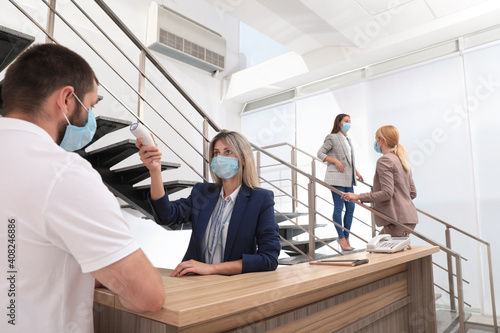 Woman in mask measuring temperature of employee with noncontact thermometer at office reception photo