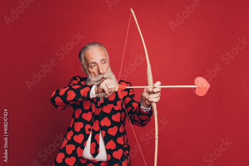 Profile photo of concentrated pensioner archer hold bow arrow aim wear heart print tux isolated red background