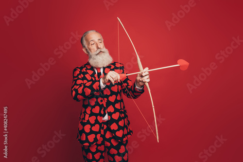 Profile photo of calm focused cupid archer old man hold bow arrow aim wear heart print suit isolated red background
