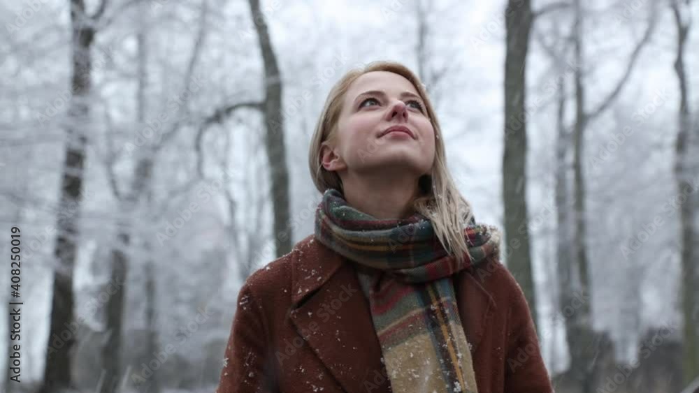 Blonde woman in scarf and coat stay in a park in snowfall Blonde woman in scarf and coat stay in a park in snowfall