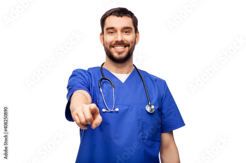 healthcare, profession and medicine concept - happy smiling doctor or male nurse in blue uniform with stethoscope pointing finger to camera over white background