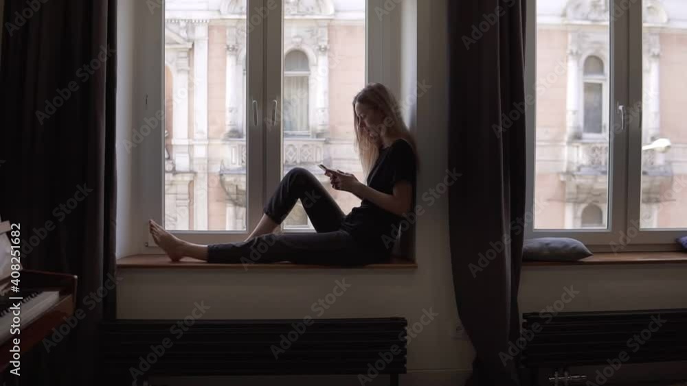 Blonde woman sitting on window sill and using smartphone in living room with piano Blonde woman sitting on window sill and using smartphone in living room with piano