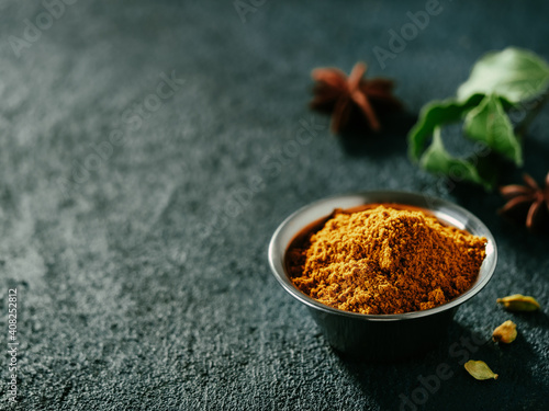 Fototapeta Naklejka Na Ścianę i Meble -  Indian or pakistani masala powder and spices on dark textured background. Small metall bowl with dry curry garam masala mix spices blend, green bay leaf, cardamom and star anise with copy space left