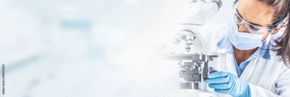 Female lab technician in protective glasses, gloves and face mask sits next to a microscope and conical flask, looking aside on the desk. - obrazy, fototapety, plakaty 