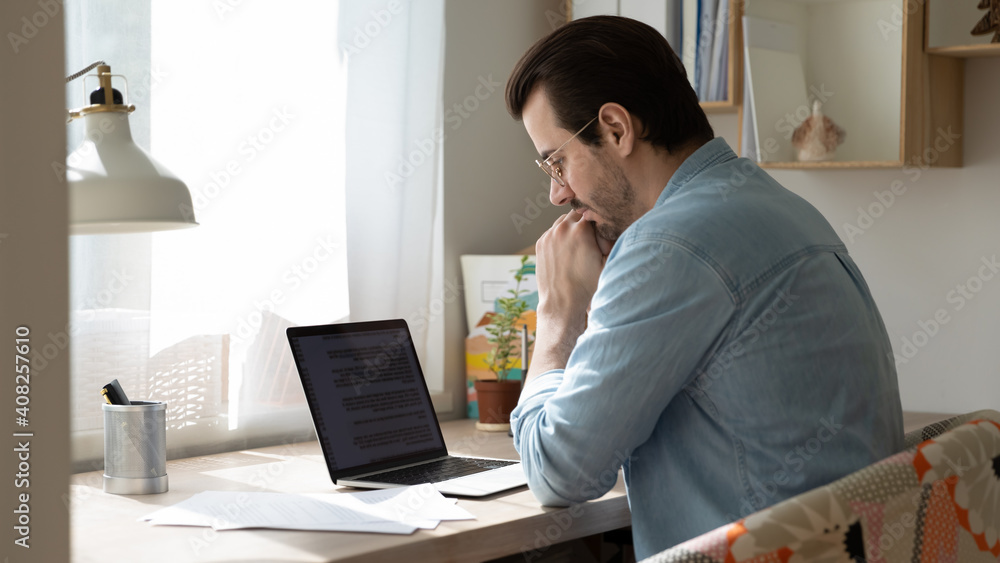 Thinking on text. Thoughtful young man working on document online or pondering on translation of electronic book in foreign language. Concentrated focused male author creating story using laptop app Thinking on text. Thoughtful young man working on document online or pondering on translation of electronic book in foreign language. Concentrated focused male author creating story using laptop app