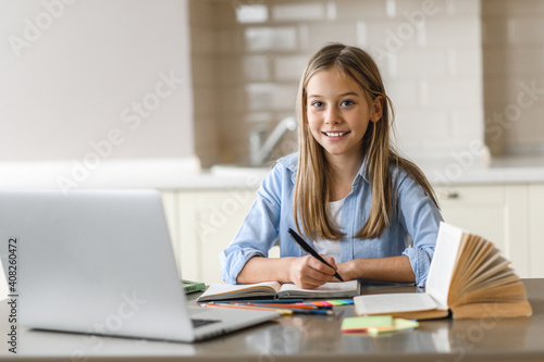 Caucasian pretty blonde schoolgirl is learning at home using laptop, sitting at the work desk, looks at the camera and smiling. Education during quarantine