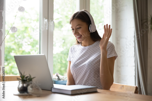 Remote classes. Smiling young female teacher tutor starting distant lesson raising hand welcoming pupils. Interested woman student sitting by pc in headphones beginning work greeting professor trainer photo