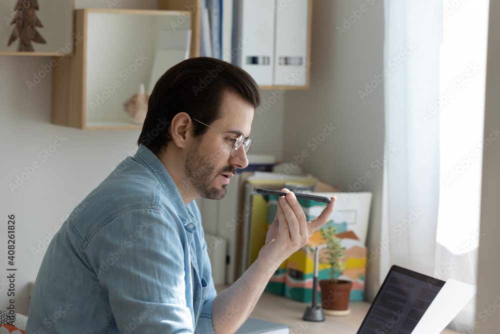 Concentrated young businessman speak to cell microphone working from home with paper electronic documents. Millennial male dictate notes by voice use phone recorder activate virtual siri assistant Concentrated young businessman speak to cell microphone working from home with paper electronic documents. Millennial male dictate notes by voice use phone recorder activate virtual siri assistant