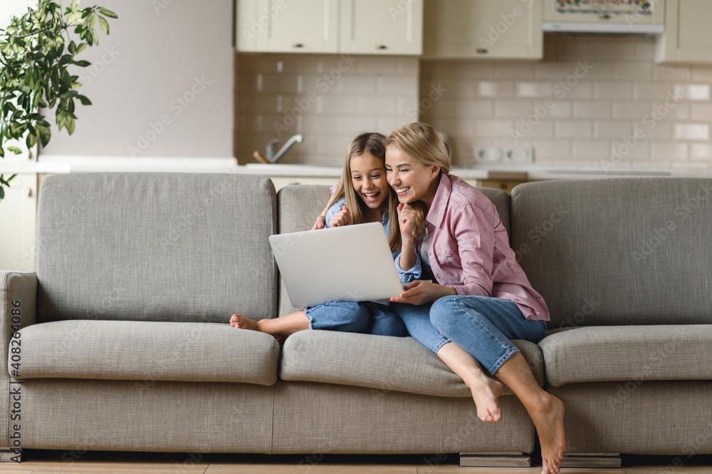 Mom and daughter spend time together at home. Happy caucasian mom and daughter are sitting on the couch, using a laptop, laughing, browsing the Internet, shopping online, seeing the good news