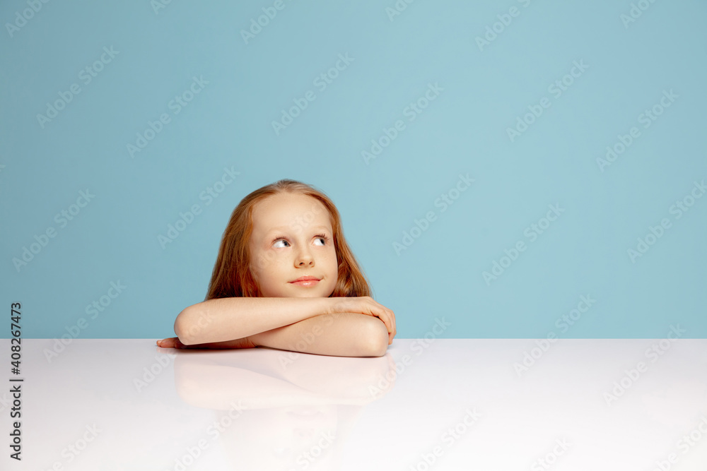 Dreamful. Happy redhair girl isolated on blue studio background. Looks happy, cheerful. Copyspace for ad. Childhood, education, emotions, facial expression concept. Sitting at the table Dreamful. Happy redhair girl isolated on blue studio background. Looks happy, cheerful. Copyspace for ad. Childhood, education, emotions, facial expression concept. Sitting at the table