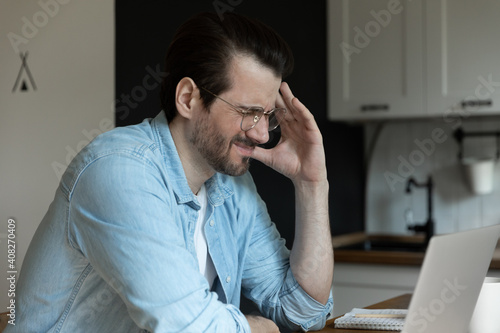 Frustrated millennial male freelancer sitting at kitchen before laptop firmly shutting his eyes unable to believe he made wrong decision. Tired young student overworked by pc feeling sudden headache