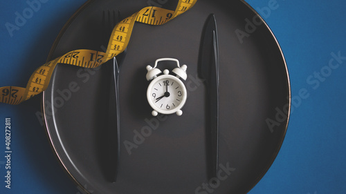 Flat lay fork and kitchen knife on a plate with tape measurement and white alarm clock. Intermittent fasting concept dieting healthy lifestyle