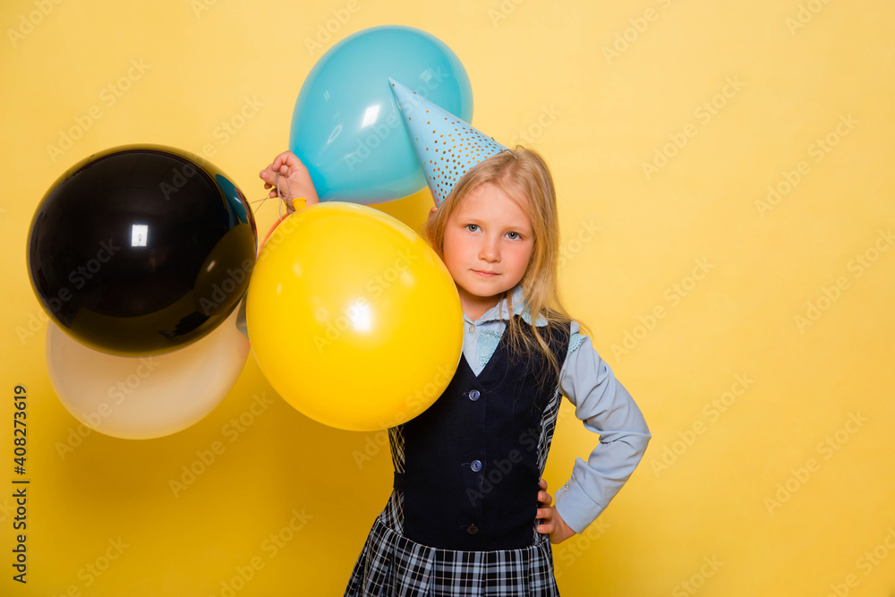 Obraz premium A girl in a school uniform with a festive cap on her head is holding balloons in her hands isolated on a yellow background.