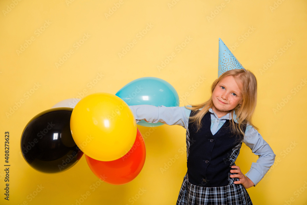 Fototapeta premium A girl in a school uniform with a festive cap on her head is holding balloons in her hands isolated on a yellow background.