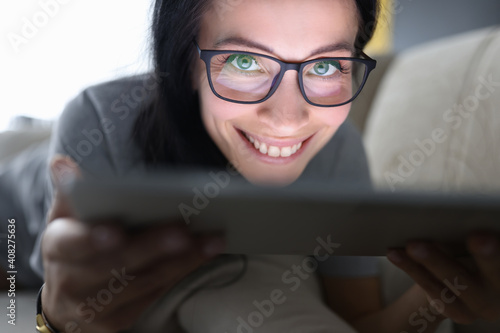 Smiling woman with glasses lies on couch and holds tablet
