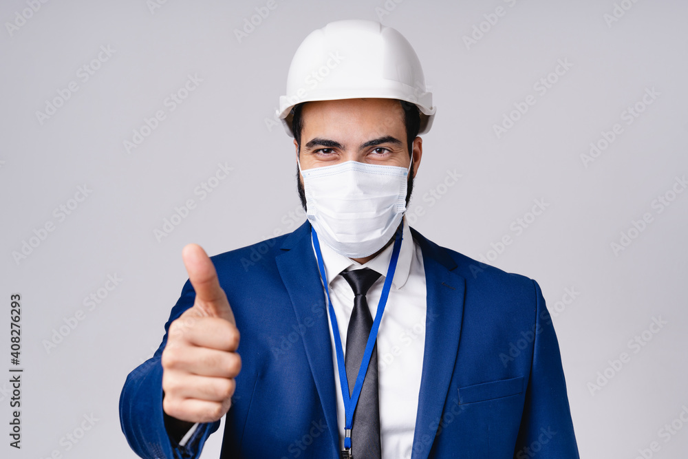 Confident young architect in hardhat and medical mask against Covid19 showing thumb up isolated over grey background Confident young architect in hardhat and medical mask against Covid19 showing thumb up isolated over grey background