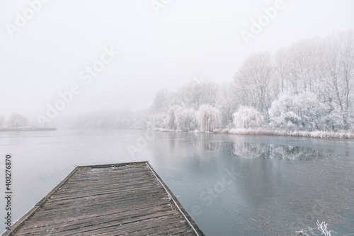 Frozen lake in winter, with wooden pier, and trees covered by snow, beautiful foggy sunset on the forest. Beautiful seasonal winter landscape, peaceful nature scenery