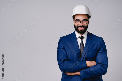 Confident young Middle Eastern egineer in white hardhat and formal suit with his arms crossed isolated over grey background photo