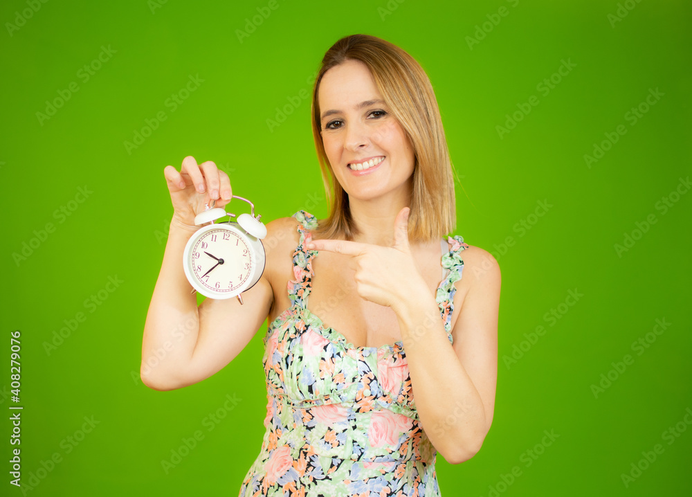 Young woman holding alarm clock standing over isolated green background very happy pointing with hand and finger Young woman holding alarm clock standing over isolated green background very happy pointing with hand and finger