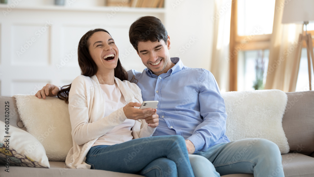 Happy young couple using phone, laughing, having fun with gadget together, sitting on cozy couch at home, overjoyed woman holding smartphone, shopping or chatting online with man, spending weekend