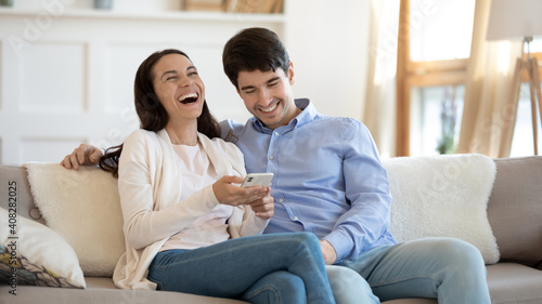 Happy young couple using phone, laughing, having fun with gadget together, sitting on cozy couch at home, overjoyed woman holding smartphone, shopping or chatting online with man, spending weekend