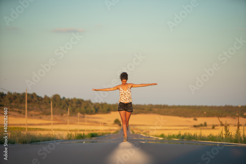 Young girl, walking in the middle of a lonely straight road, late in the day. With the hands stretched out in the shape of a cross, to maintain balance and not fall from the dividing line.