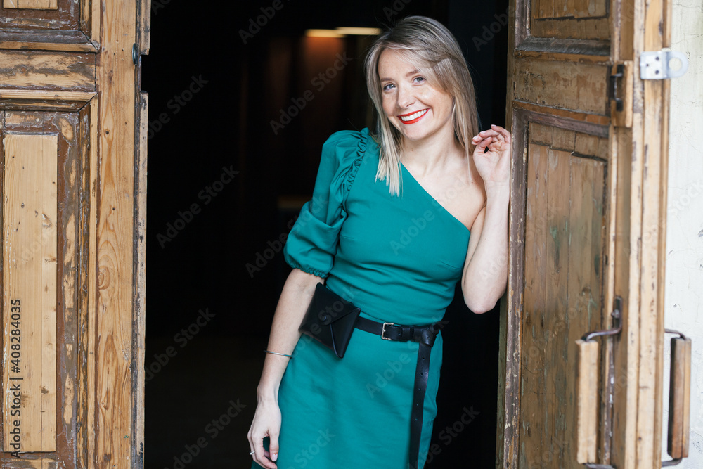 beautiful young smiling blonde woman with red lipstick and in a green dress stands at the exit from the building between the old wooden doors beautiful young smiling blonde woman with red lipstick and in a green dress stands at the exit from the building between the old wooden doors