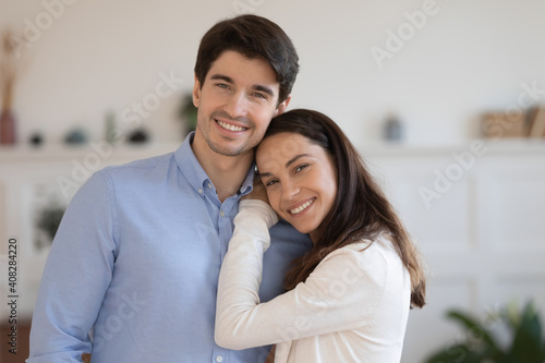 Head shot portrait smiling man and woman hugging, standing at home, happy young couple posing for family photo together, positive wife and husband looking at camera, cuddling, enjoying leisure time