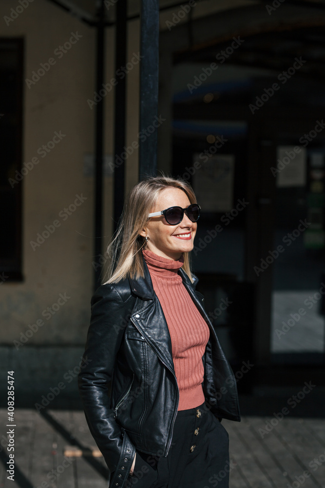 Obraz premium young beautiful smiling stylish blonde woman in sunglasses, black pants and leather jacket posing on the don of buildings in an urban setting on sunny day