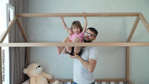 Young father with little daughter do gymnastics on horizontal bar of crib in form of house at home. Sport and gymnastics concept. Self-isolation. Father and child girl play active games.