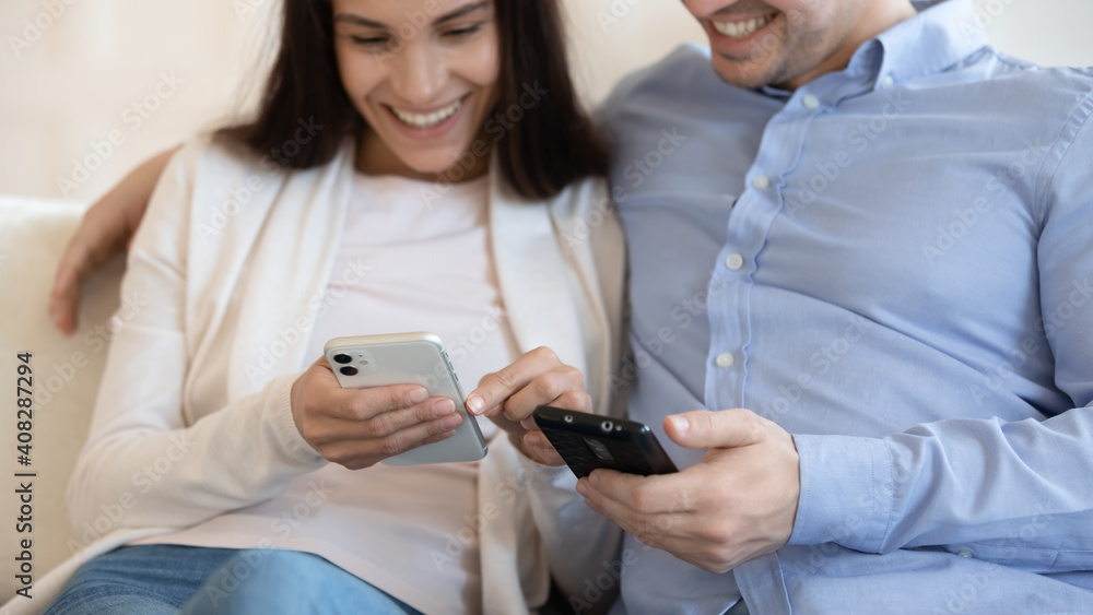 Close up smiling woman and man using phones, sharing news, hugging, sitting on couch together, young couple surfing internet, holding smartphones, having fun with gadgets, enjoying leisure time