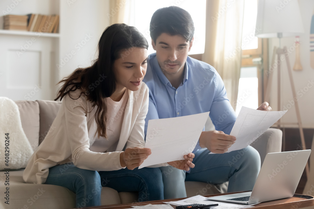 Close up serious young couple reading documents, calculating finances, sitting on couch at desk with laptop and calculator, focused wife and husband checking mortgage agreement, planning budget Close up serious young couple reading documents, calculating finances, sitting on couch at desk with laptop and calculator, focused wife and husband checking mortgage agreement, planning budget